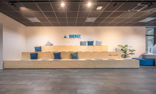Offene Empfangs- und Kommunikationszone mit großzügiger Sitztribüne, integrierter Präsentationsfläche und markentypischem BENZ-Branding im Verwaltungsgebäude der BENZ Tooling GmbH in Gengenbach. Der multifunktionale Begegnungsbereich fördert den Austausch zwischen Mitarbeitenden, Kunden und Gästen und verbindet Kommunikation, Präsentation und Unternehmenskultur in einer modernen Arbeitswelt. BENZ entwickelt und produziert hochpräzise CNC-Aggregate, Werkzeugsysteme und Komponenten für den Maschinen- und Anlagenbau sowie für die Holz-, Metall- und Verbundwerkstoffbearbeitung. Bauherr: BENZ Tooling GmbH. Geplant und umgesetzt von arc+ GmbH Freudenstadt.