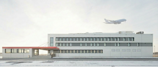 Airline-Catering- und Logistikzentrum der SCK BBI Immobilien GmbH & Co. KG in Berlin-Schönefeld mit direkter Nähe zum Flughafen und startendem Flugzeug als Symbol für internationale Logistik, kurze Wege und hochfunktionale Industriearchitektur. Bauherr: SCK BBI Immobilien GmbH & Co. KG. Ort: Berlin-Schönefeld. Geplant und umgesetzt von arc+ GmbH Freudenstadt.