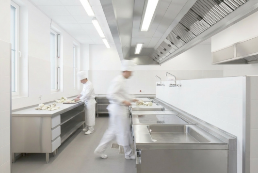 Hygienisch ausgestattete Großküche mit Edelstahlarbeitsplätzen und Mitarbeitenden in der Lebensmittelproduktion für das Airline-Catering im Logistikzentrum der SCK BBI Immobilien GmbH & Co. KG in Berlin-Schönefeld. Bauherr: SCK BBI Immobilien GmbH & Co. KG. Ort: Berlin-Schönefeld. Geplant und umgesetzt von arc+ GmbH Freudenstadt.